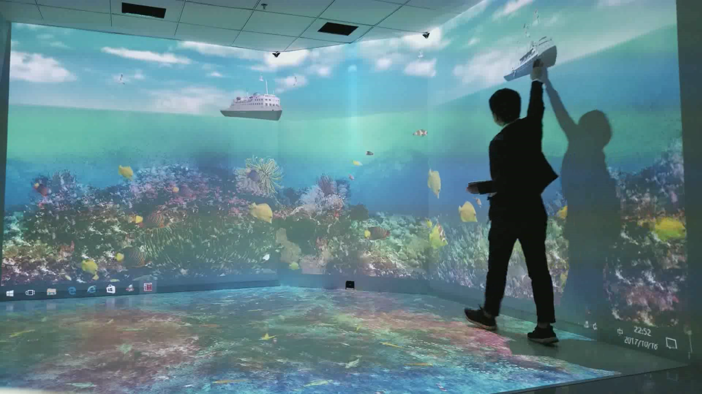 A man walks along a wall with his hand touching a projection of a boat. The walls and floor are all covered in projected ocean images.