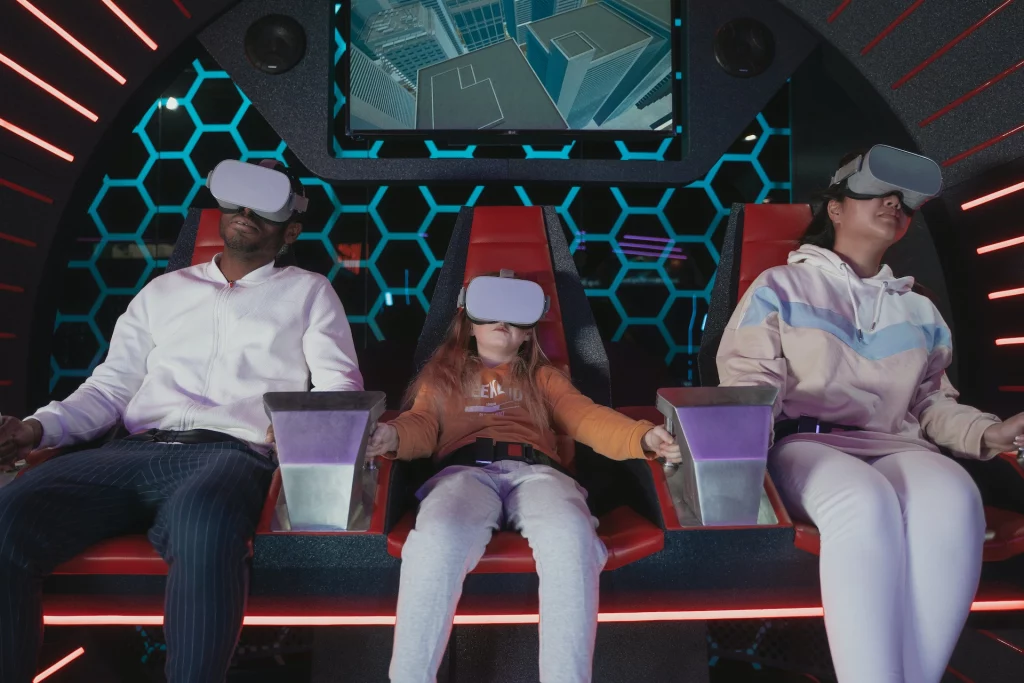 Two adults and a child wearing VR headsets sitting in special seats with a honeycomb pattern on the wall behind them and a display on the wall showing tops of buildings