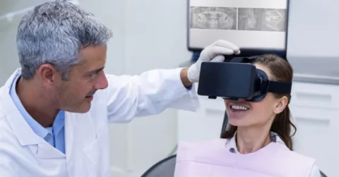 A man in a lab coat assists a woman in an exam chair with adjusting a VR headset