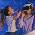 A young woman helps another young woman adjust a VR headset.