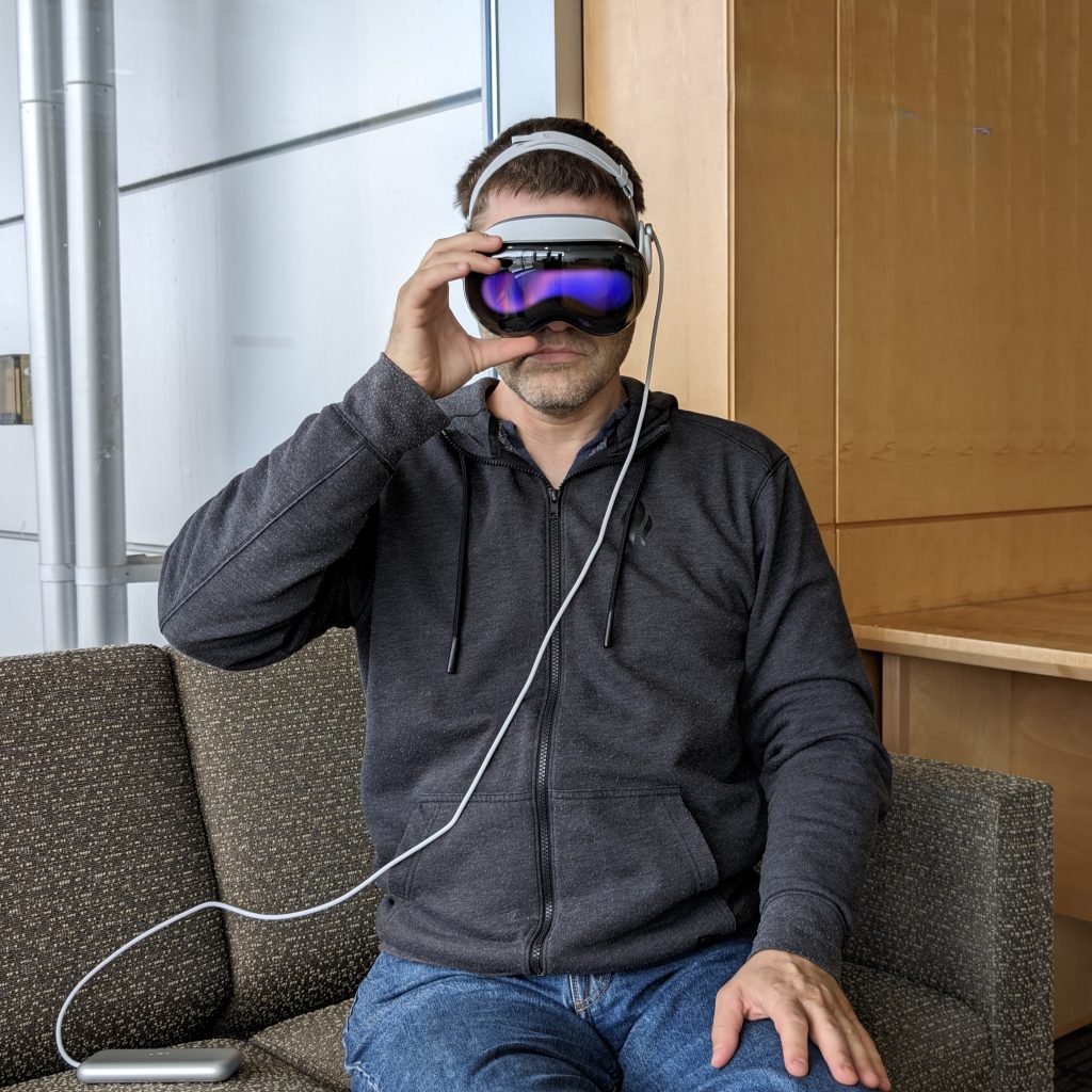A man sitting on a couch wearing jeans and a gray jacket using the Apple Vision Pro headset.